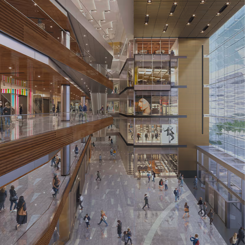 Interior view of a modern shopping mall with multiple levels, glass walls, and people walking on the ground floor and upper balconies.